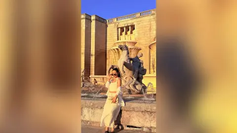 Woman in white summer outfit posing by a stone fountain statue at a historic building during golden hour.