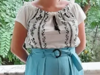 Woman in white bohemian embroidered blouse and light blue belted skirt standing in a lush garden setting.