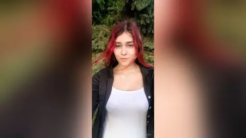 Young woman with red hair wearing a white tank top and black jacket in a lush green forest setting.