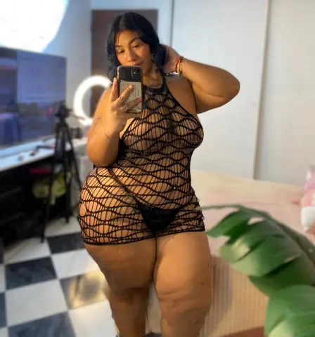 NatashaSantt — NatashaSantt poses in a black mesh net bodysuit that shows her skin through the netting, standing in a room with a ring light and bed.