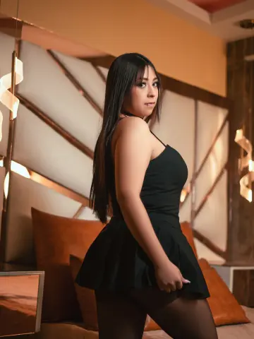 Young woman in a black cocktail dress posing in a luxury bedroom with warm ambient lighting.