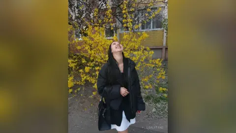 Cheerful woman in a black puffer jacket looking up at blooming yellow flowers in a spring garden.