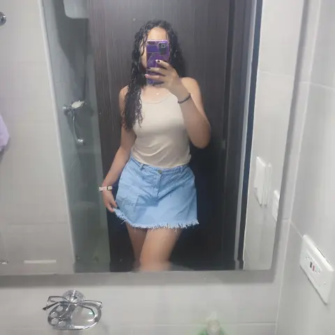 Young woman in a white tank top and light blue denim skirt taking a mirror selfie in a modern bathroom.