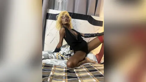 Glamorous blonde woman in a black plunging bodysuit lounging on a bed in a chic, moody setting.