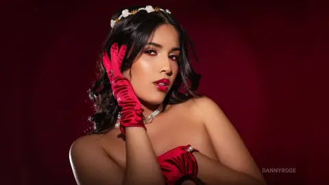 Elegant woman in red satin gloves and pearl headband posing against a moody dark red background.