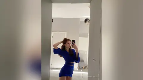 Young woman in a royal blue satin outfit taking a mirror selfie in a bright, modern minimalist room.