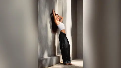 Young woman in white tank top and baggy black jeans leaning against a concrete wall in moody sunlight.
