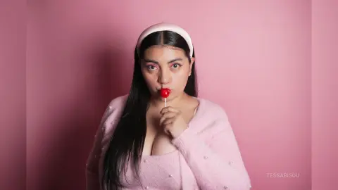 Young woman in a pink fuzzy sweater and headband eating a red lollipop against a pastel pink background.