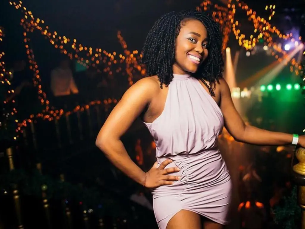TeelaAdams smiles in a tight, light purple halter dress with a high slit, posing in a dark club setting with warm string lights.