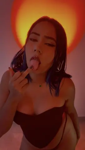 Young woman in a black bodysuit posing with a playful expression against a warm orange sunset glow.