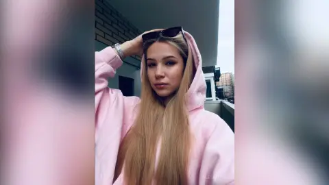 Aletakunde wears a pink hoodie with her long blonde hair hanging down while she holds sunglasses on her head outdoors.