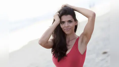 Smiling brunette woman in a red swimsuit posing on a sunny beach with long wavy hair.