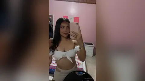 Young woman in a white tie-front crop top taking a selfie in a pink bedroom.