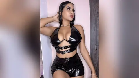 MiaKorz poses in a black shiny halter top and matching tight shorts, showing off her cleavage and curves against a plain wall.