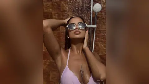 Glamorous woman in a lavender bikini and white sunglasses posing under a shower in a rustic outdoor setting.