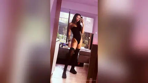 LucyRosenfelds poses in a black bodysuit and black thigh-high stockings, standing in a room with a large window behind her.