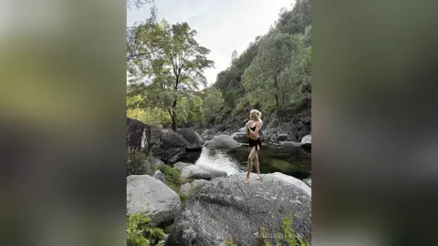 Blonde woman in a black swimsuit standing on rocks by a serene forest river in a peaceful natural setting.