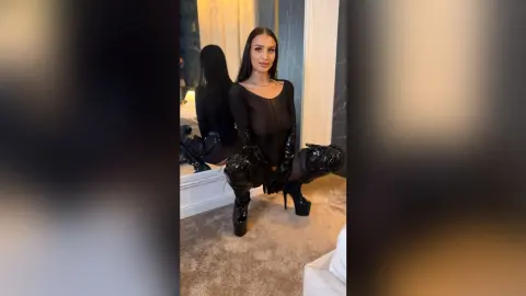 LeyaGrifin poses in a black bodysuit and thigh-high patent leather boots while squatting in a room.