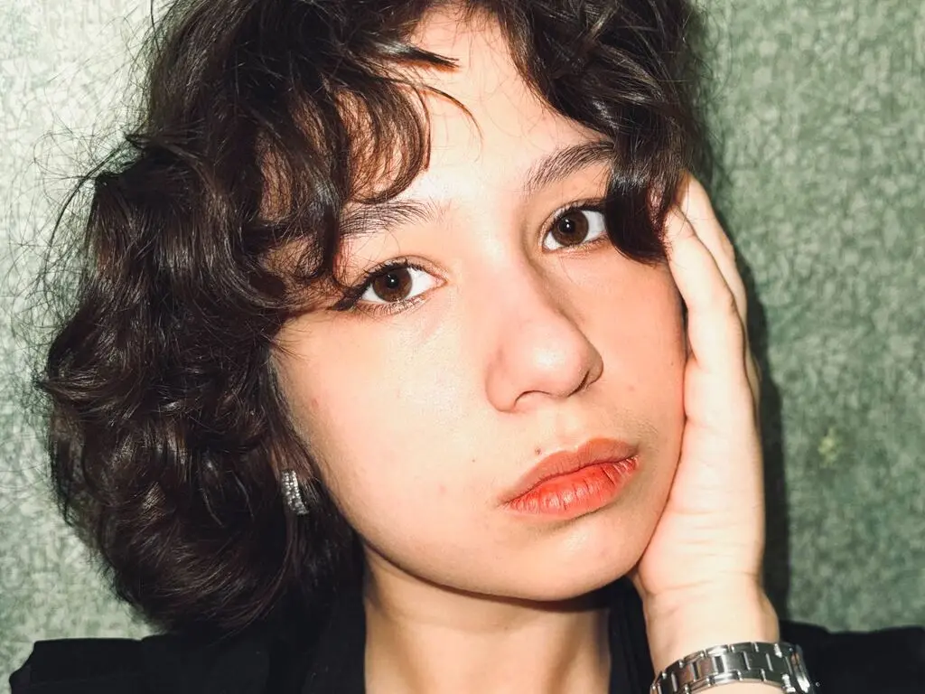 Close-up portrait of a young woman with wavy brown hair posing against a textured green wall in soft lighting.