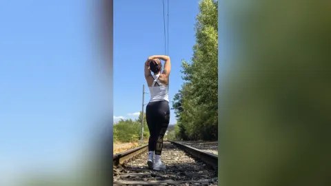 JaneHope — JaneHope stands on railroad tracks in a white tank top and black leggings with her hands behind her head.