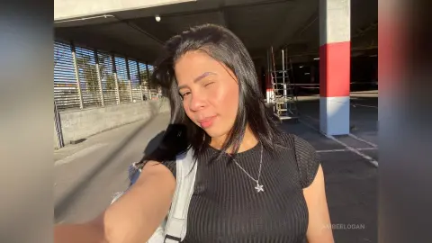 Smiling woman in a black ribbed top and silver star necklace posing in a sunny outdoor parking garage.
