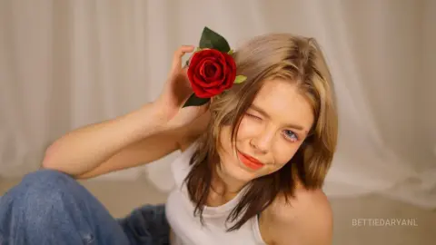 Smiling young woman in a white tank top holding a red rose near her face against a neutral backdrop.