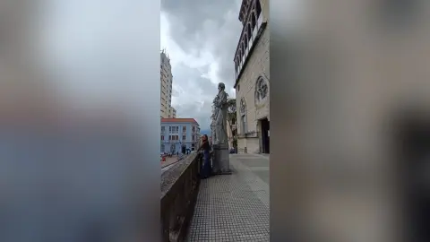 AmelieLautrec stands outdoors next to a stone statue and a low wall, wearing a dark top and blue jeans against a city building background.
