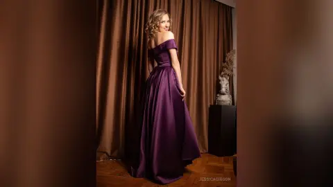 Smiling woman in an elegant off-the-shoulder purple satin gown standing against brown curtains in a studio.