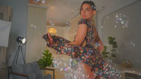 Woman in a floral summer dress playing with bubbles in a bright, dreamy indoor setting.