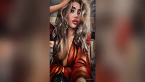 Glamorous woman in a red satin wrap top posing with wavy blonde hair in a moody indoor setting.