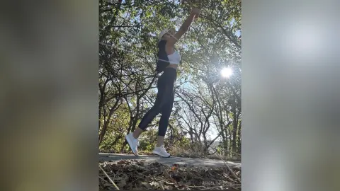 Woman in white crop top and black leggings stretching outdoors in a sunny forest setting.