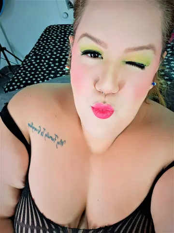 Woman with green eyeshadow and pink lips wearing a black tank top in a cozy indoor setting.