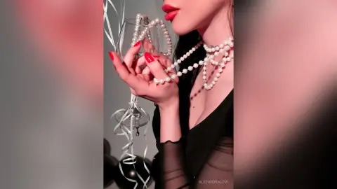 Elegant woman in a black dress and pearl necklace holding a crystal glass against a soft pink background.
