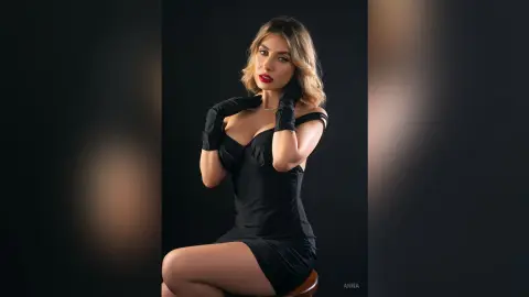 Elegant woman in a black cocktail dress and gloves posing glamorously against a dark studio background.