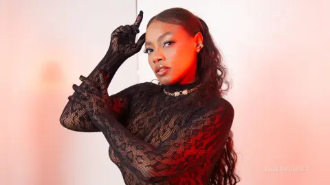 Glamorous woman in a black lace turtleneck bodysuit posing against a warm red studio background.