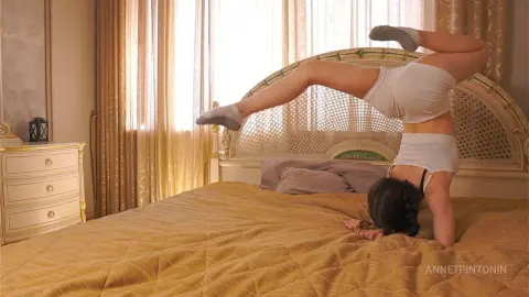 Young woman in white loungewear doing a yoga stretch on a bed in a sunlit, cozy bedroom.