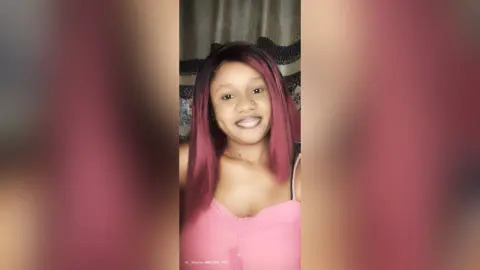 KiraDon smiles while wearing a pink top with her long, reddish-pink hair styled straight down.