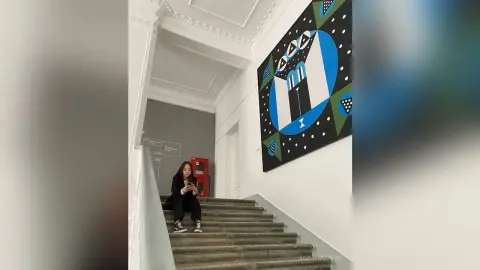 Young woman in black outfit sitting on white stairs next to large modern abstract art in a bright hallway.