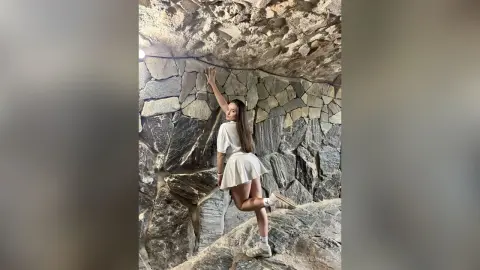 Young woman in a white summer outfit posing against a rustic stone wall in a moody, natural setting.