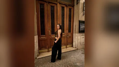 Stylish woman in black crop top and sequin trousers standing before ornate wooden doors in a moody urban setting.