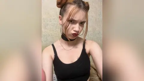 Young woman with space buns and dark lipstick wearing a black tank top in a minimalist indoor setting.