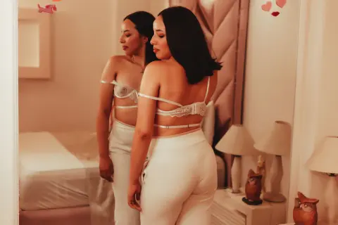 Elegant woman in white lace lingerie and trousers posing in a warm, cozy bedroom setting.