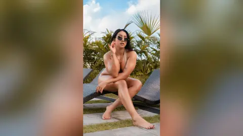 KimRoberth sits on a black lounge chair wearing sunglasses and a white bikini, posing with her legs crossed outdoors.