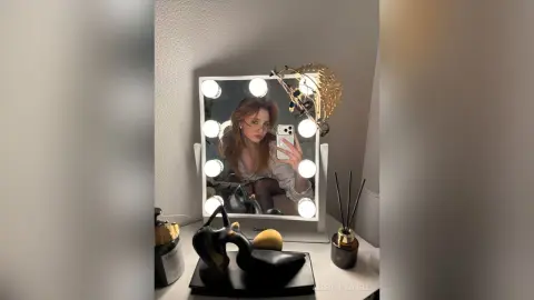 AlbertaCorrell takes a mirror selfie in her makeup chair, wearing glasses and a light blouse.