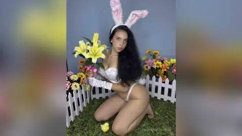Young woman in white bunny ears and lingerie kneeling in a spring garden setting with yellow lilies.