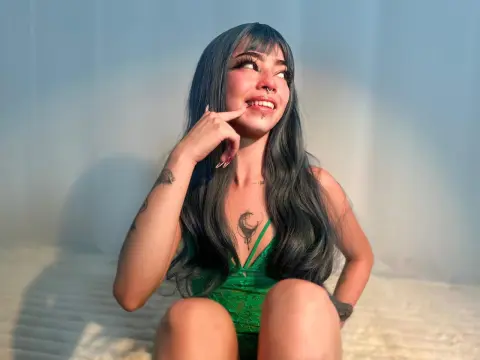Smiling young woman with long dark hair and tattoos wearing a green lace bodysuit in a bright studio setting.