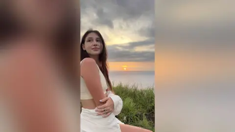 RitaMoonllight poses outdoors at sunset, wearing a white crop top and white skirt with her long brown hair down.