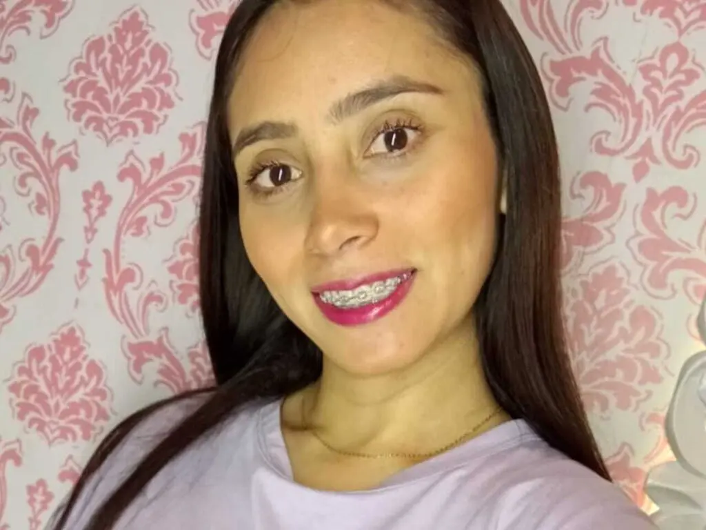 Smiling young woman with long dark hair and braces wearing a lavender t-shirt against a pink damask wallpaper.