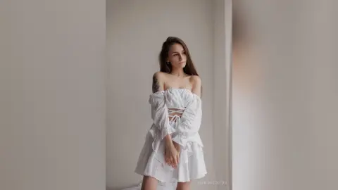 IngridBeckton poses in a white off-the-shoulder lace dress with long sleeves, looking away while standing against a plain white wall.