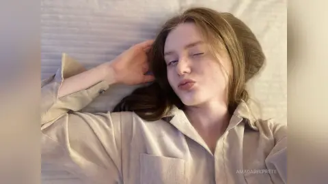 Young woman in a beige linen shirt lying on a white bed with a relaxed, dreamy expression.
